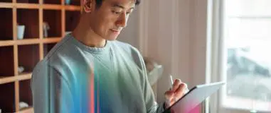 Image of a man in a casual grey sweatshirt using a stylus to work on a tablet, focused and engaged, in a well-lit indoor setting with shelves and a window in the background.