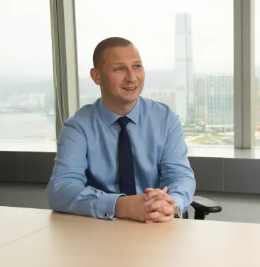 青いシャツを着たビジネスマンがデスクに座り、都市の景色を背景に笑顔を見せている様子